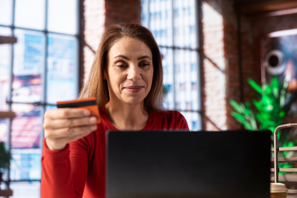 Uśmiechnięta kobieta w czerwonym swetrze trzymająca kartę płatniczą przed laptopem, symbolizująca udaną konwersję w sklepie internetowym.